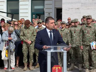 Un bărbat într-un costum închis și cravată stă în spatele unui pupitru transparent, în fața unui grup de persoane îmbrăcate în uniforme militare.