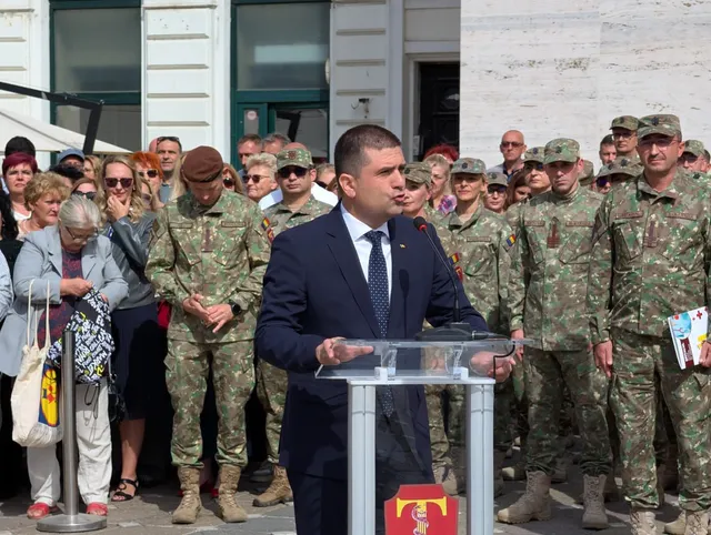 Un bărbat într-un costum închis și cravată stă în spatele unui pupitru transparent, în fața unui grup de persoane îmbrăcate în uniforme militare.