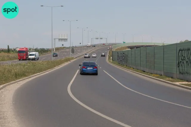 Un autoturism albastru circulă pe o autostradă cu mai multe benzi, lângă un gard înalt și un camion roșu parcat pe marginea drumului.