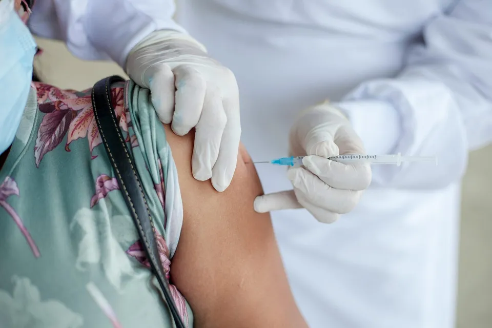 O persoană cu o cămașă cu model floral primește o injecție în braț de la o persoană îmbrăcată în alb și purtând mănuși medicale albe.