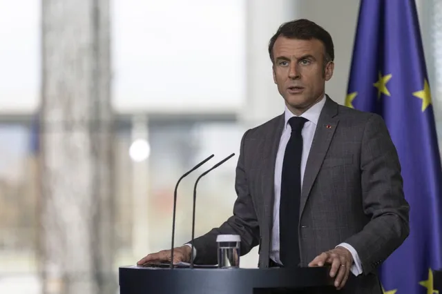 Un bărbat într-un costum gri și cravată neagră stă în spatele unui podium cu două microfoane, lângă un steag albastru cu stele galbene.