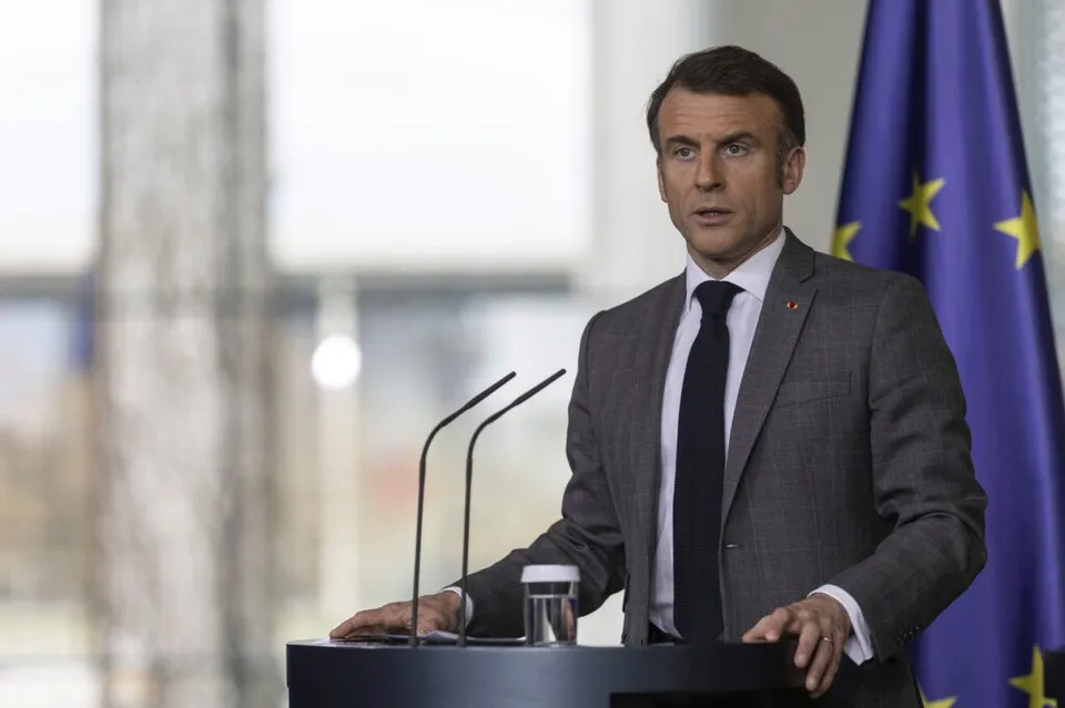 Un bărbat într-un costum gri și cravată neagră stă în spatele unui podium cu două microfoane, lângă un steag albastru cu stele galbene.