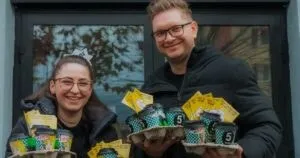 Un bărbat și o femeie cu ochelari țin în mâini tăvi cu pahare de cafea și pachete de produse galbene în fața unei ferestre.