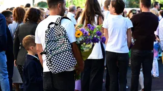 Un grup de persoane stă în picioare, printre care un bărbat cu un rucsac alb și negru și o femeie care ține un buchet de flori.