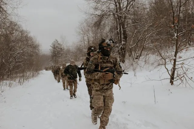 Soldați în uniforme de camuflaj și echipament tactic merg printr-un peisaj de iarnă acoperit de zăpadă printre copaci fără frunze.