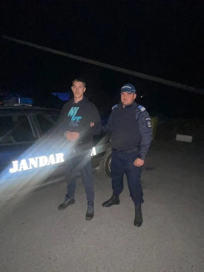 Un bărbat într-un hanorac negru și un agent de jandarm în uniformă albastră stau lângă un vehicul de serviciu noaptea.