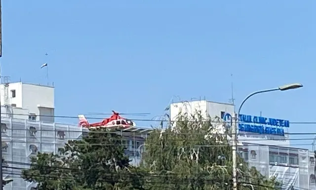 Un elicopter roșu este poziționat deasupra unei structuri de clădire albe sub un cer senin.