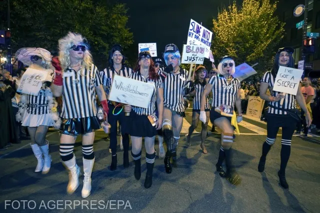 Un grup de persoane poartă costume cu dungi verticale albe și negre, accesorii de cap și ține diverse panouri de carton în timpul unei procesiuni nocturne.