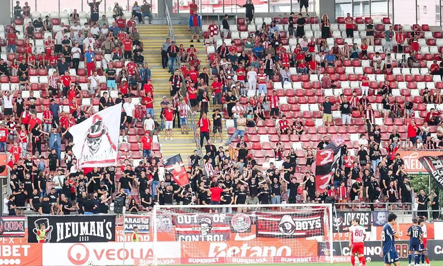 Mulți oameni stau pe tribunele unui stadion cu scaune roșii și albe, purtând haine de culoare roșie și neagră.