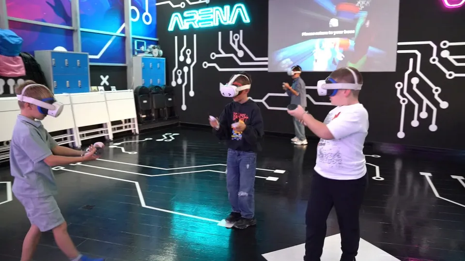 Trei copii poartă căști de realitate virtuală și țin controllere în timp ce se află într-o încăpere cu podea neagră și linii albe.