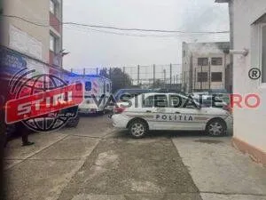 O mașină de poliție albă și o ambulanță sunt parcate pe un drum asfaltat lângă clădiri și un gard de sârmă.