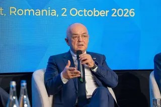 Un bărbat cu ochelari și costum albastru stă pe un scaun și vorbește într-un microfon în fața unui ecran albastru.