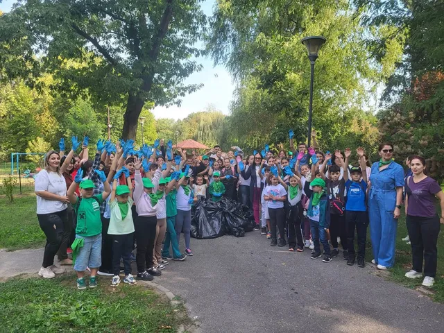 Un grup mare de oameni stă pe un drum asfaltat, mulți purtând mănuși de protecție colorate și ridicând mâinile în aer lângă saci negri de gunoi.