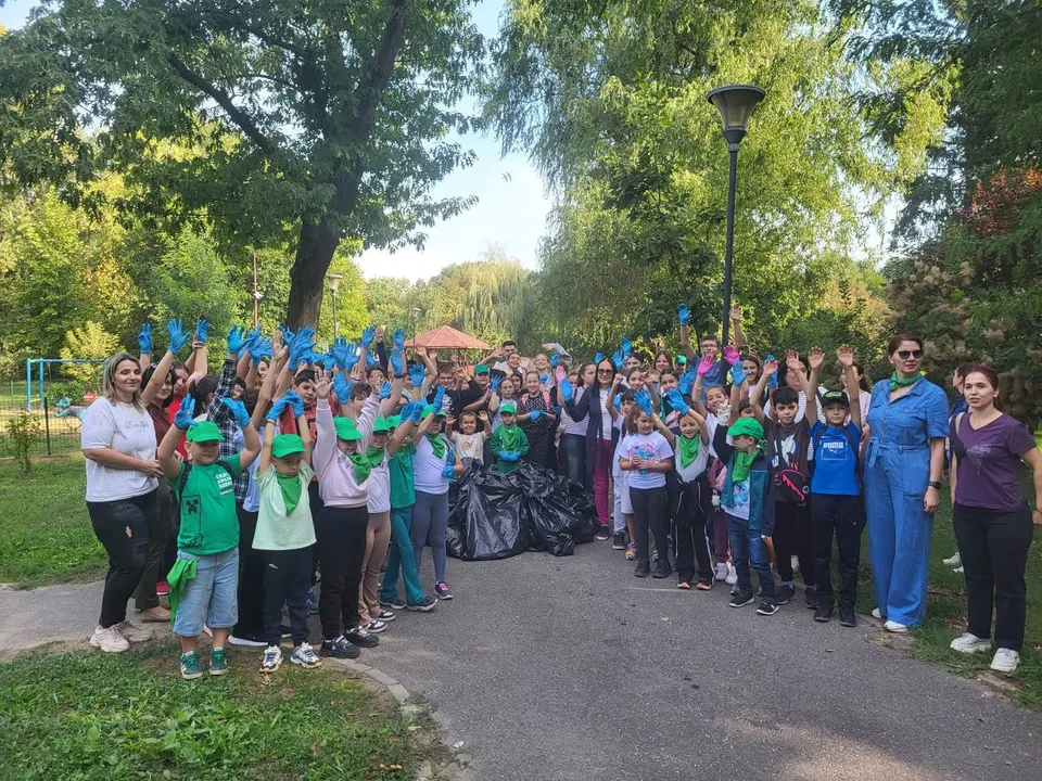 Un grup mare de oameni stă pe un drum asfaltat, mulți purtând mănuși de protecție colorate și ridicând mâinile în aer lângă saci negri de gunoi.