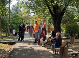 Un grup de persoane stă pe o cărare pietonă în parc, lângă un copac mare și bănci de lemn sub lumina soarelui.