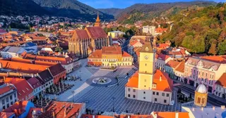 O vedere aeriană a unui oraș cu clădiri cu acoperișuri roșii, o catedrală înaltă și o piață centrală pavată lângă dealuri împădurite.