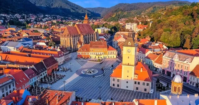 O vedere aeriană a unui oraș cu clădiri cu acoperișuri roșii, o catedrală înaltă și o piață centrală pavată lângă dealuri împădurite.