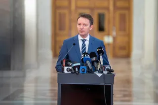 Un bărbat într-un costum albastru și cravată striptiză stă în spatele unui podium pe care sunt amplasate mai multe microfoane.