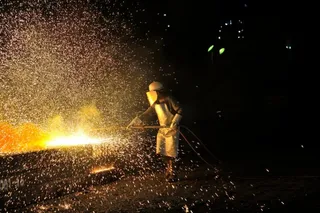 O persoană purtând echipament de protecție și mască de sudură manipulează un instrument de lângă o sursă de scântei strălucitoare pe fundal negru.