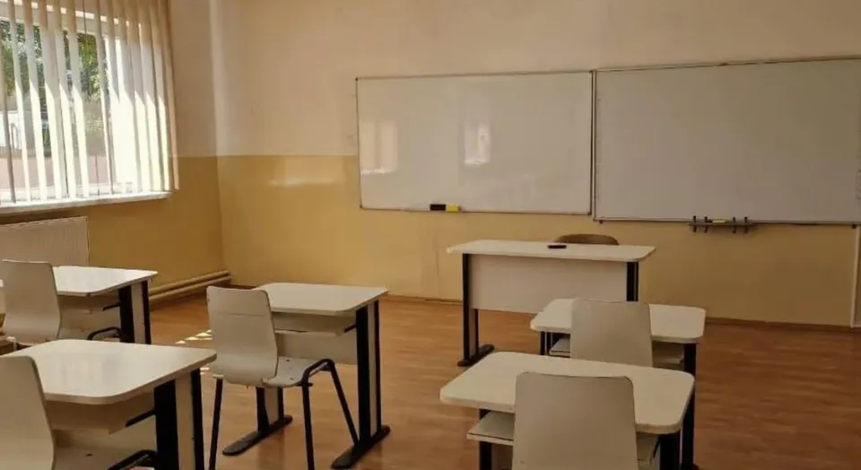 O sală de clasă cu mai multe birouri albe și scaune gri dispuse pe un podea de lemn în fața a două table albe mari.