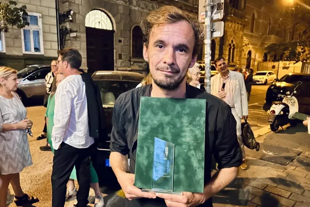 Un bărbat cu părul ciufulit ține un obiect dreptunghiular verde și un trofeu transparent în fața unui grup de persoane pe o stradă noaptea.