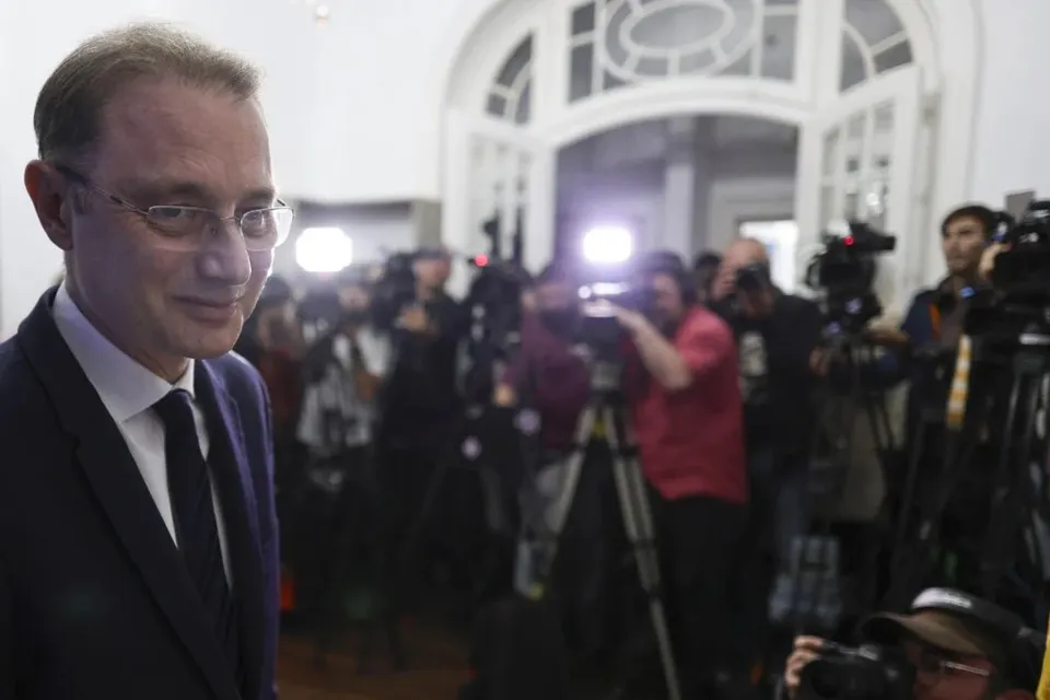 Un bărbat cu ochelari și costum în prim-plan privește spre lateral, în timp ce un grup de fotografi și cameramani cu echipamente se află în fundal.