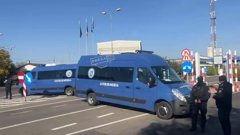 Două autoutovalite albastre cu inscripția JANARMERIA sunt parcate pe un drum asfaltat lângă doi bărbați în uniforme negre sub un cer senin.