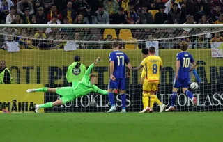 Un fotbalist într-o echipă verde este întins pe teren în fața porții, în timp ce alți jucători în echipamente albastre și galbene privesc mingea.