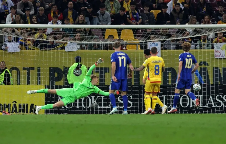 Un fotbalist într-o echipă verde este întins pe teren în fața porții, în timp ce alți jucători în echipamente albastre și galbene privesc mingea.