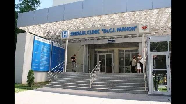 O intrare de spital cu scări, un panou informativ albastru și câteva persoane stând lângă ușa de sticlă a clădirii.