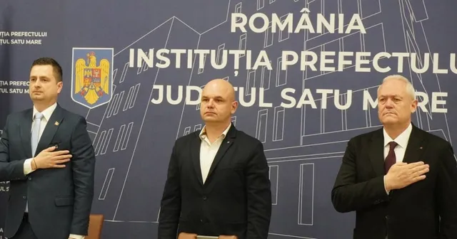Trei bărbați stau în picioare în fața unui panou albastru pe care scrie text alb și un blazon colorat.