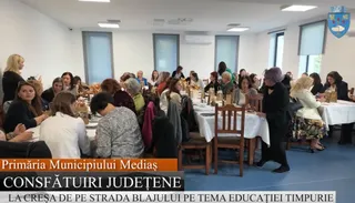 Un grup de persoane stă așezat la mese cu fețe albe într-o încăpere iluminată, participând la un eveniment cu text scris pe un afiș de jos.