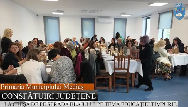 Un grup de persoane stă așezat la mese cu fețe albe într-o încăpere iluminată, participând la un eveniment cu text scris pe un afiș de jos.