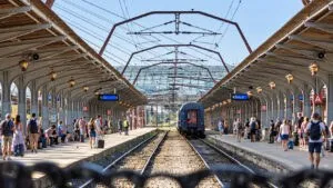Un tren gri stă pe un drum feroviar între două platforme de gară acoperite, unde persoane stau pe peron.
