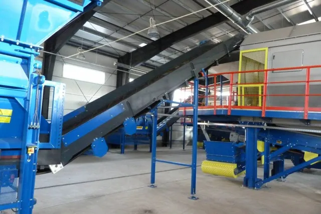 O instalație industrială pentru sortarea deșeurilor cu o bandă transportoare înclinată, echipamente albastre și o platformă cu balustradă roșie.