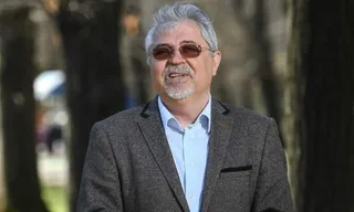 Un bărbat cu părul și barba gri, purtând ochelari și un sacou maro peste o cămașă albastră, stă în fața unor copaci.