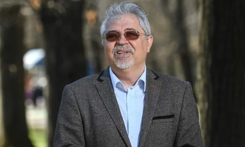 Un bărbat cu părul și barba gri, purtând ochelari și un sacou maro peste o cămașă albastră, stă în fața unor copaci.