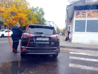 Un bărbat în haine închise stă lângă partea din spate a unui autoturism negru parcat pe marginea unei străzi lângă o trecere de pietoni.