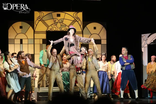 Un grup de actori în costume de epocă pozează pe o scenă de teatru în fața unui decor cu elemente geometrice și arcuri doricești.