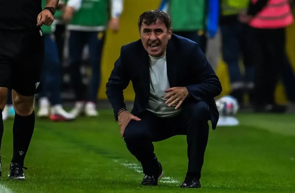 Un bărbat cu sacou închis și tricou deschis se ghemuiește pe un teren de fotbal verde, având o expresie tensionată.