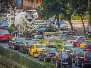 O mulțime de mașini colorate și un mixer de beton sunt parcate pe o stradă lângă un trotuar cu copaci verzi.
