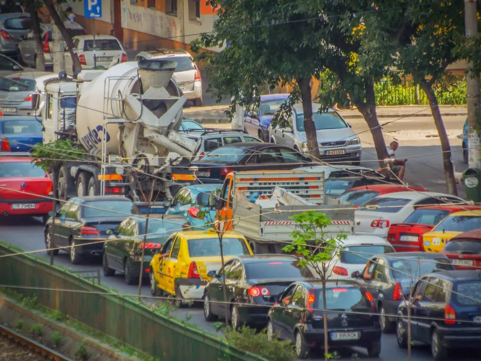 O mulțime de mașini colorate și un mixer de beton sunt parcate pe o stradă lângă un trotuar cu copaci verzi.