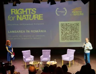 Un bărbat cu microfon și o femeie stau pe o scenă lângă trei scaune de culoarea lavandei și un ecran mare cu text și cod QR.