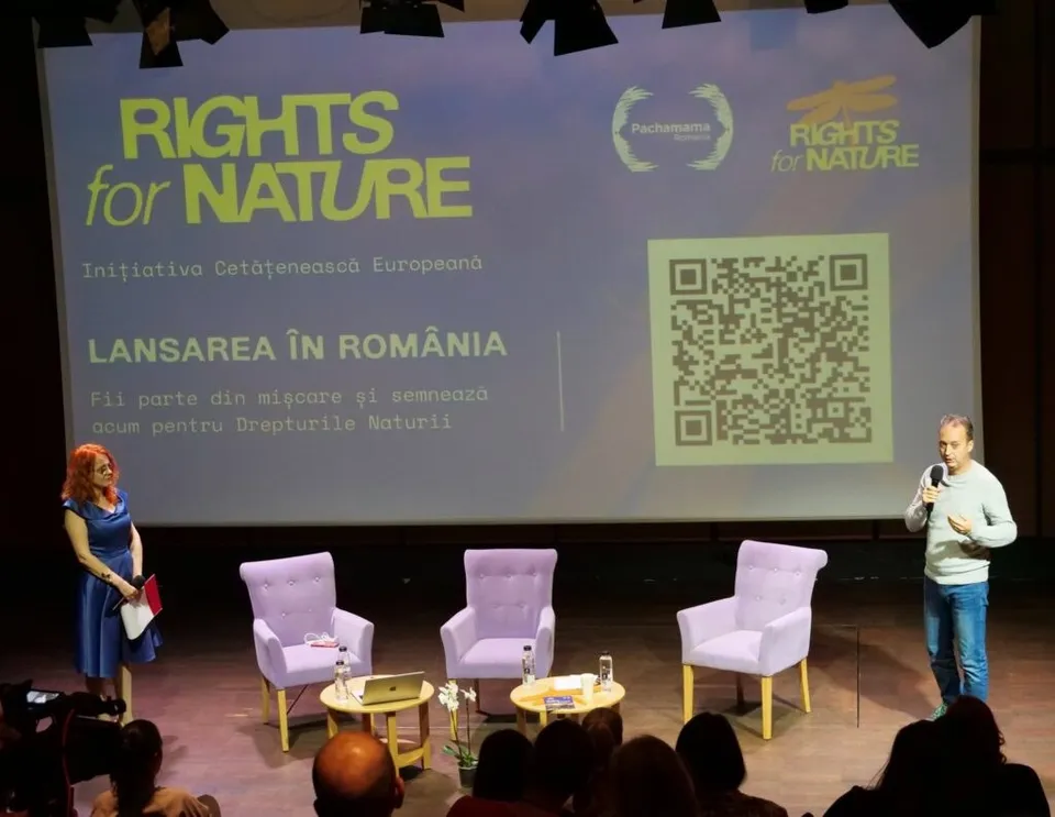 Un bărbat cu microfon și o femeie stau pe o scenă lângă trei scaune de culoarea lavandei și un ecran mare cu text și cod QR.