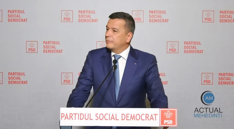 Un bărbat cu costum albastru și cămașă albă stă în fața unui fundal cu logoul partidului PSD repetat de mai multe ori.