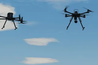 Două drone multicopter negre zboară în cerul albastru cu nori albiștri dispersi, echipate cu antene și camere de filmat.
