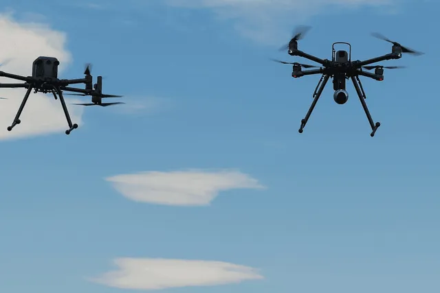 Două drone multicopter negre zboară în cerul albastru cu nori albiștri dispersi, echipate cu antene și camere de filmat.