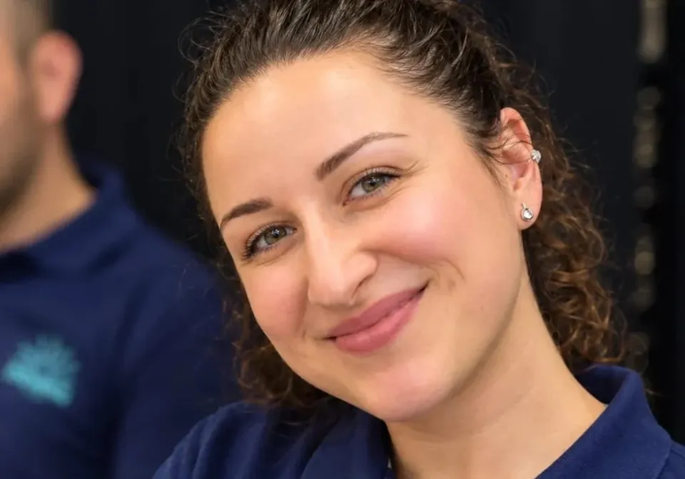 O femeie cu păr castaniu ondulat, prins în spate, poartă o bluză bleumarie cu guler și zâmbește în prim-plan.