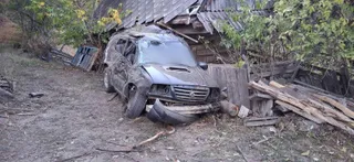 Un autoturism de culoare închisă, avariat, este poziționat lângă o construcție din lemn și stive de scânduri într-o zonă cu vegetație.