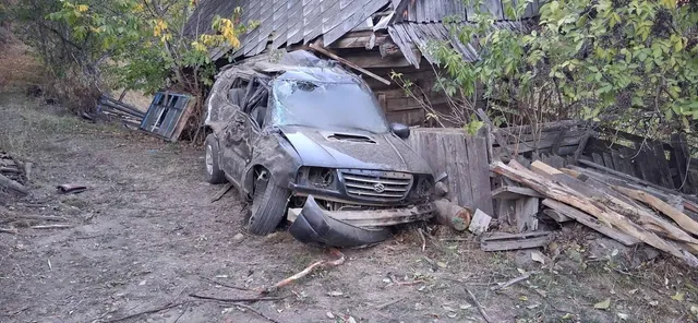 Un autoturism de culoare închisă, avariat, este poziționat lângă o construcție din lemn și stive de scânduri într-o zonă cu vegetație.